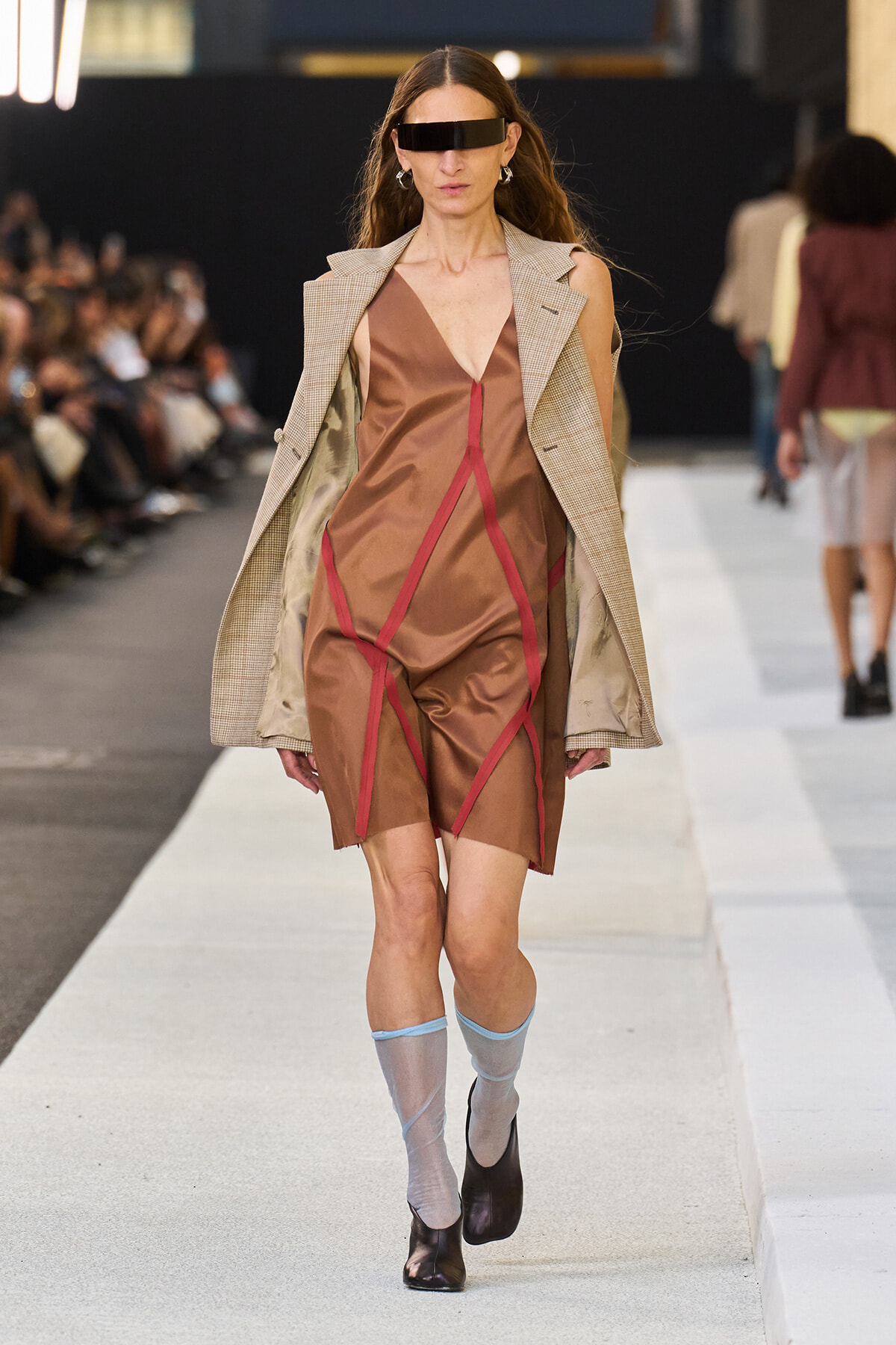 Model on a runway wearing a brown V-neck dress with red seam piping, a beige plaid blazer draped over her shoulders, and light blue sheer socks with brown ankle boots.