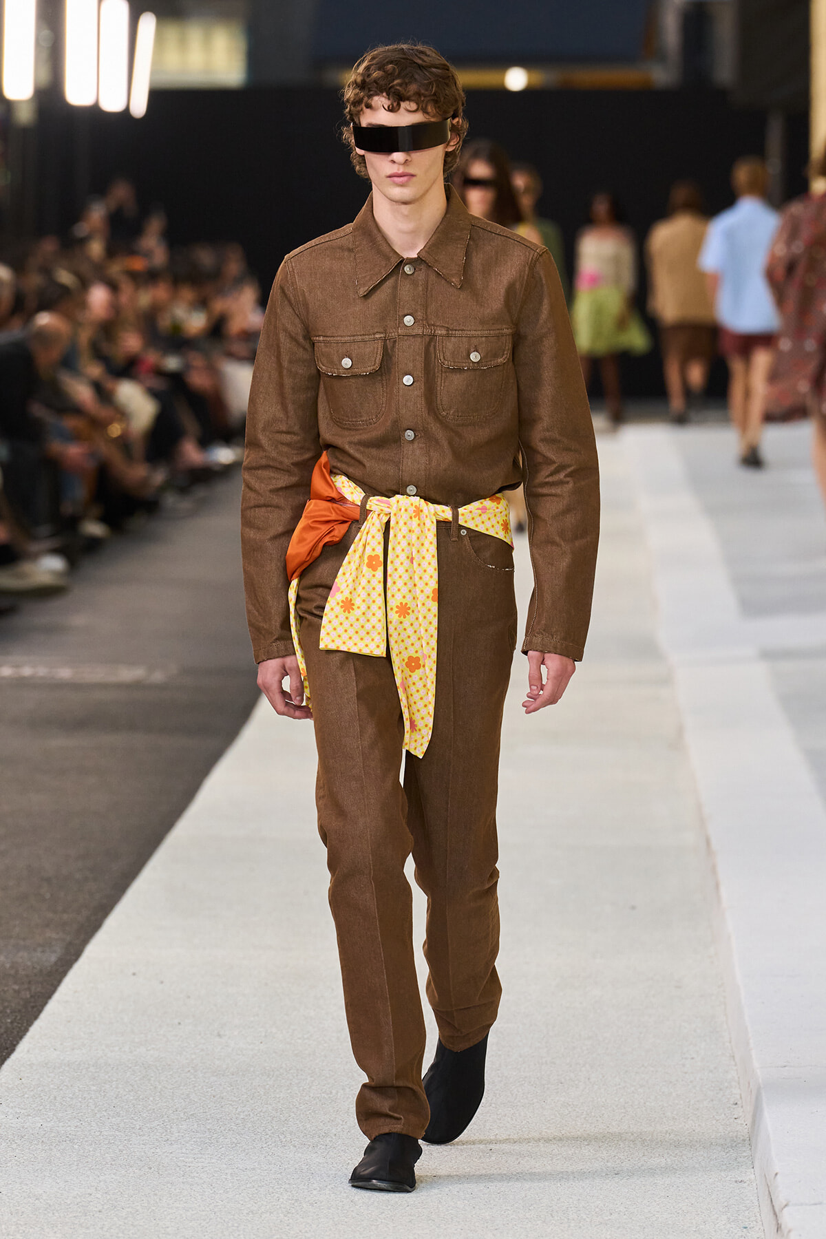 Male model walking a runway in a brown denim jumpsuit with front pockets and a yellow floral belt scarf tied at the waist.