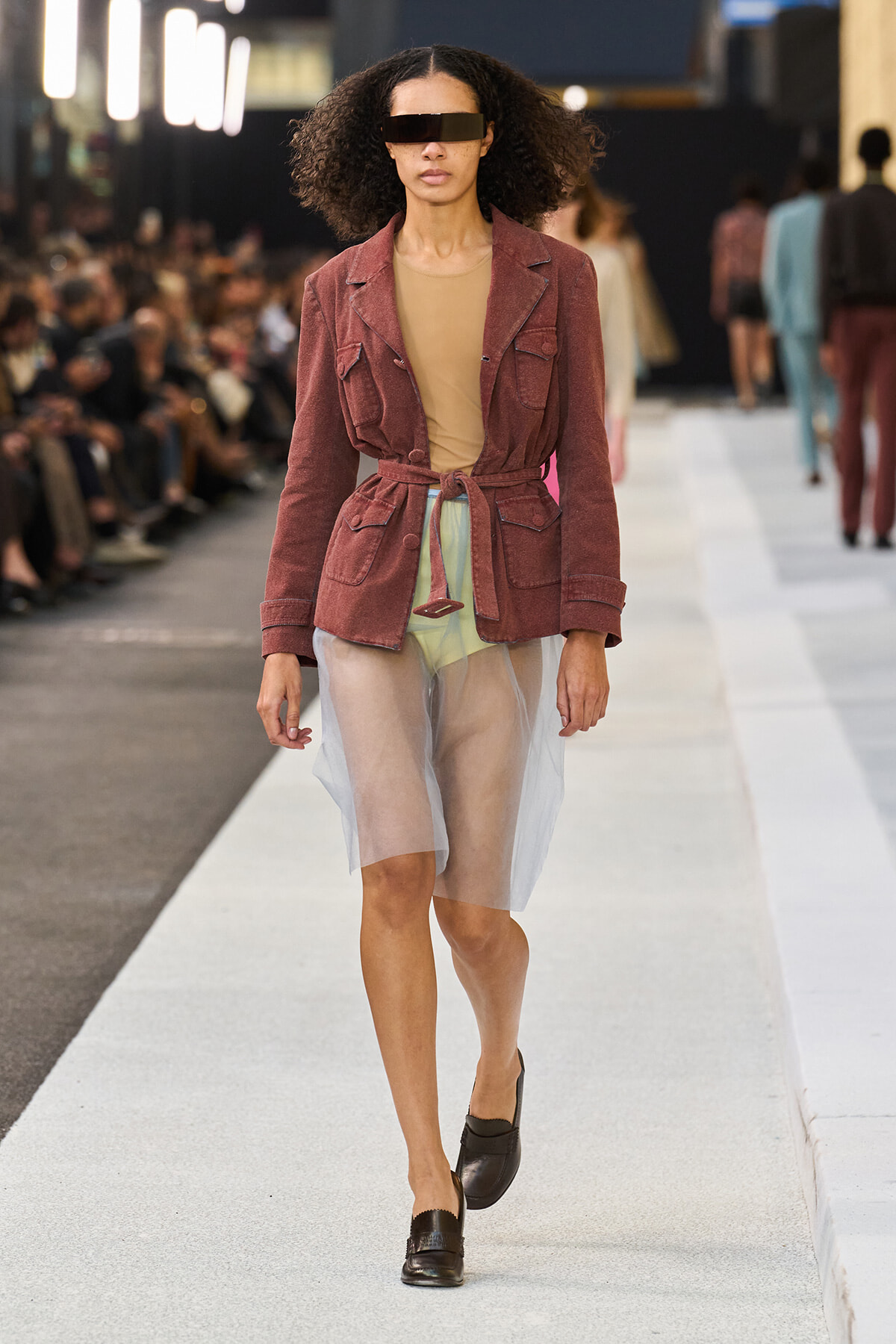 Model walks runway in a burgundy belted utility jacket with a sheer white skirt and tan top, wearing dark sunglasses.