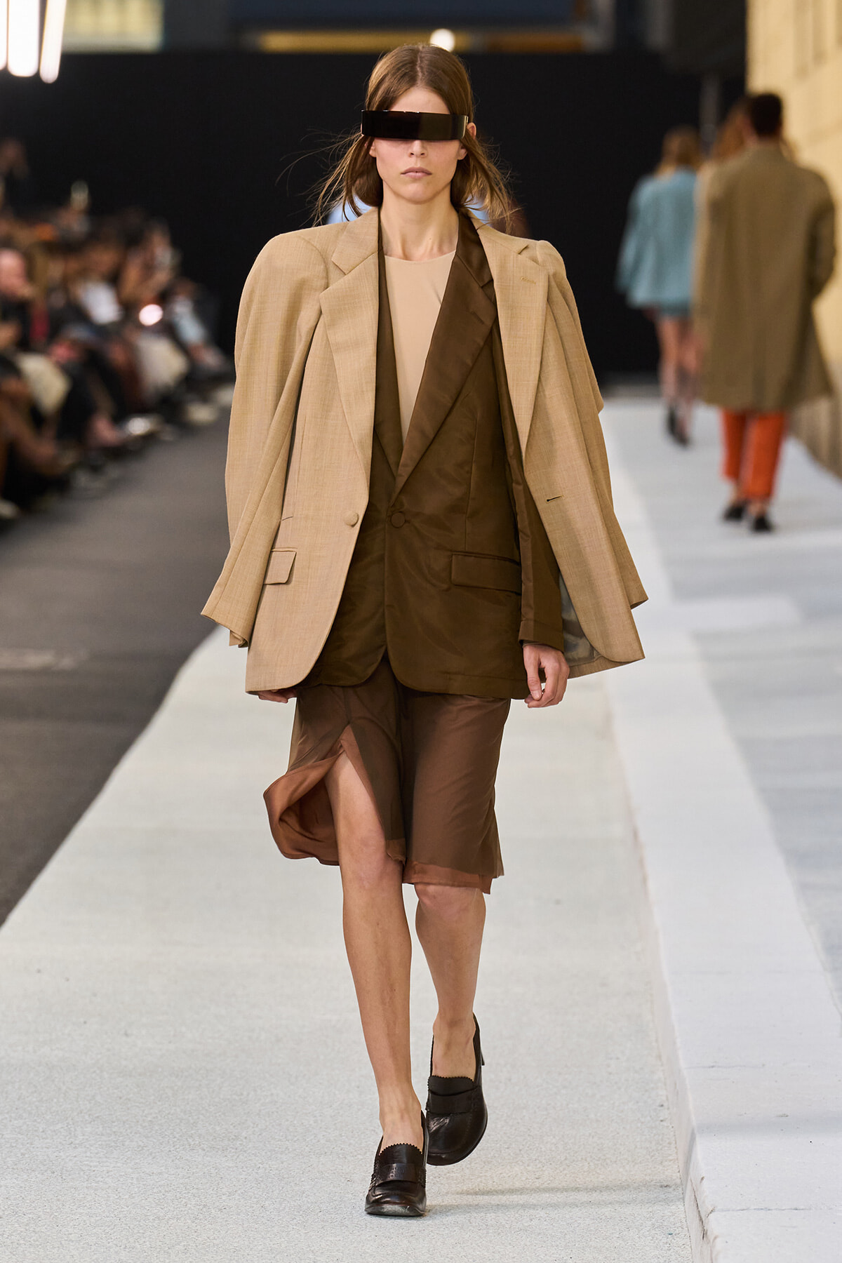 Runway model wearing a beige oversized blazer over a brown suit with a knee-high slit skirt and black rectangular visor sunglasses.