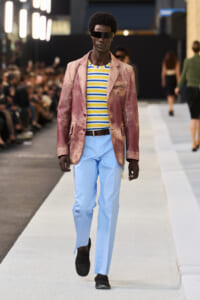 Male model on a runway wearing a pink velvet blazer, yellow-blue striped shirt, and light blue trousers with a black belt and black shoes.