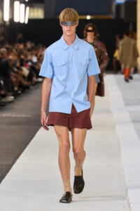 Male model on a runway in a light blue short-sleeve shirt with pockets, burgundy shorts, beige socks, black loafers, and a metallic eye visor.
