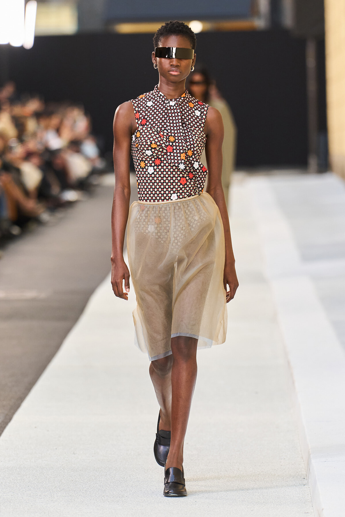 Model walking a fashion runway in a sleeveless floral top, sheer beige knee-length skirt, black visor sunglasses, and black loafers.