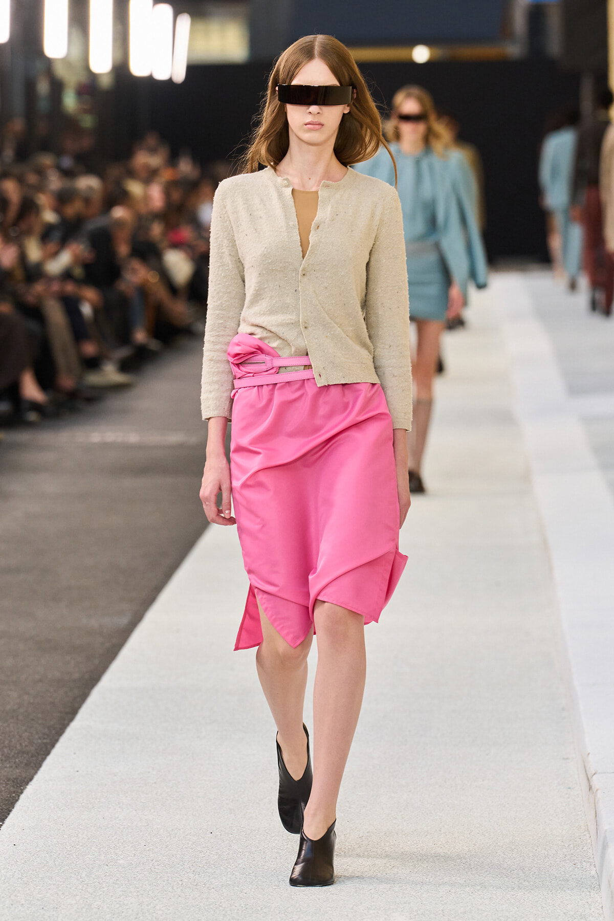 Model walks a fashion runway in a cream cardigan and bright pink satin skirt, with black ankle boots and futuristic black visor sunglasses.