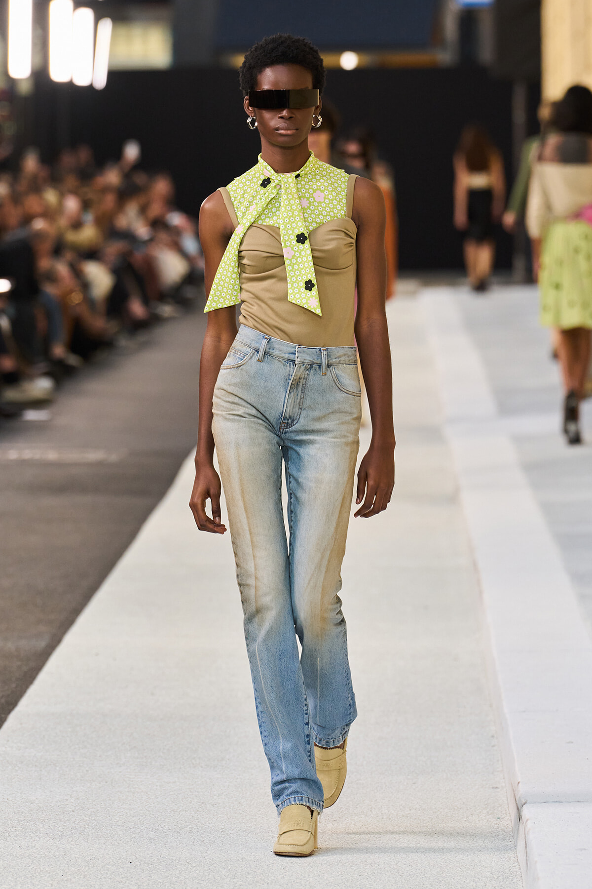 Runway model walking the catwalk in a lime-green patterned scarf tied at the neck over a beige bustier, faded jeans, and yellow boots, with dark sunglasses.