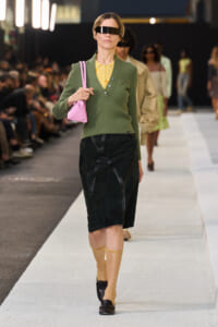 Model on a runway in a green polo over a yellow blouse, black pencil skirt, tan socks, and loafers, carrying a pink handbag with a futuristic visor headset.