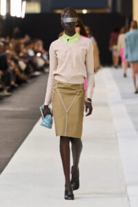 Model walking a fashion runway in a beige cardigan, lime patterned shirt collar, tan pencil skirt with decorative seams, blue clutch, and a metallic eye visor.