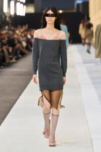 Model walking a runway in a dark gray off-the-shoulder sweater dress with a colorful strapless bikini top showing, beige leg warmers, and pink sandals with fringe; futuristic visor sunglasses cover eyes.