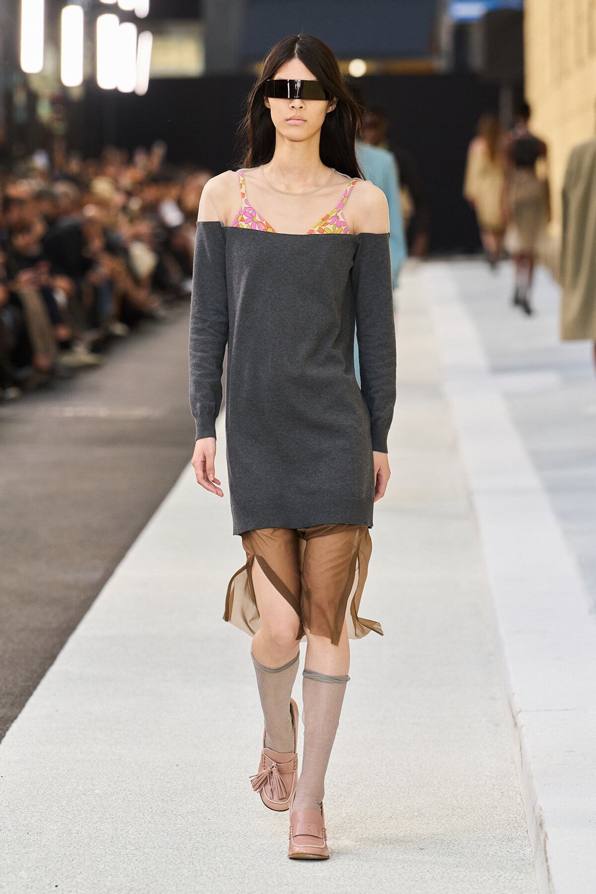 Model walking a runway in a dark gray off-the-shoulder sweater dress with a colorful strapless bikini top showing, beige leg warmers, and pink sandals with fringe; futuristic visor sunglasses cover eyes.