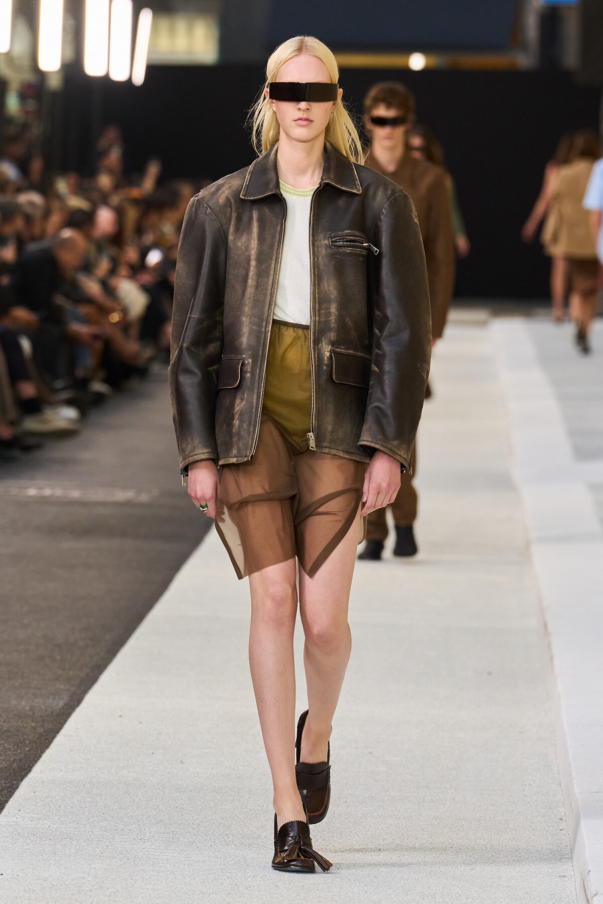 Model walks the runway in a distressed brown leather jacket, tan shorts, and a black rectangular eye visor; audience in the background.