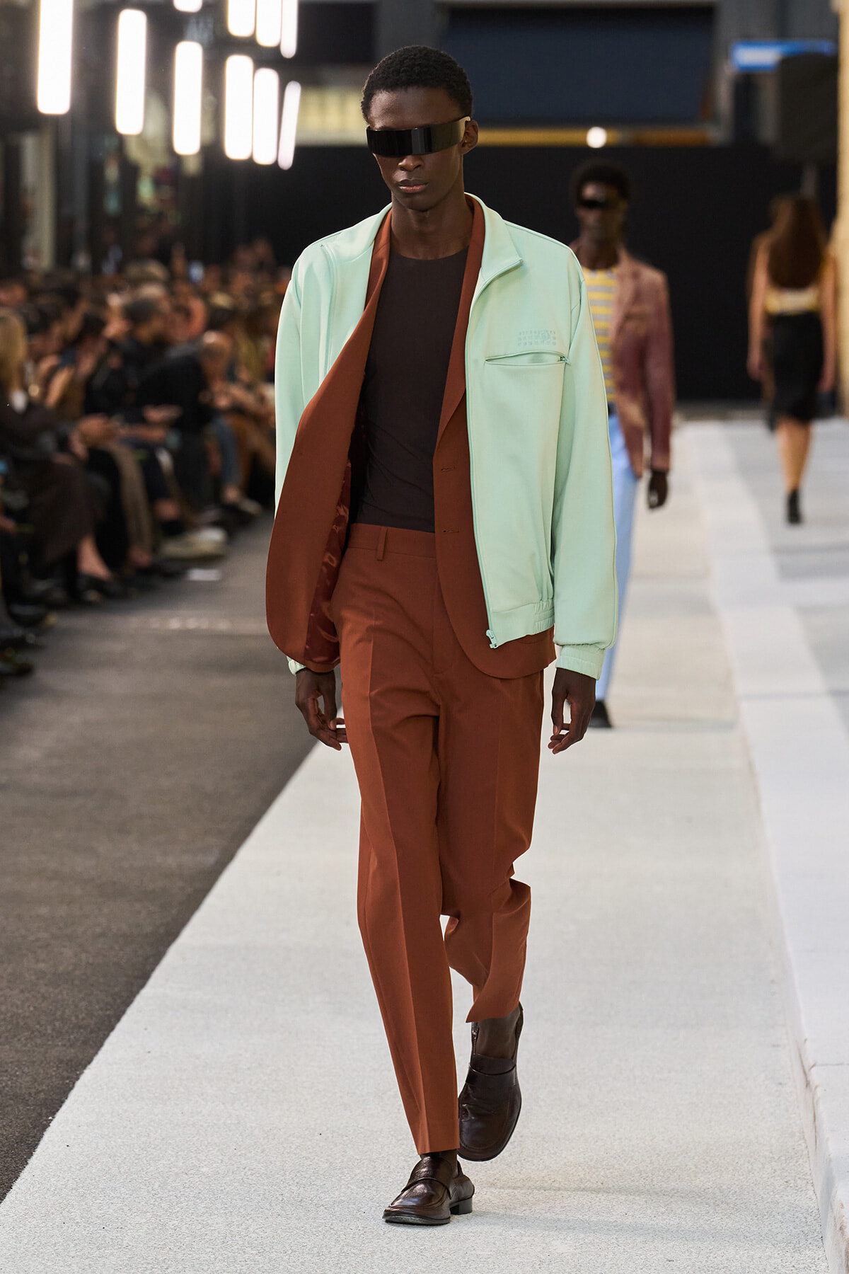 Male model walking a fashion show runway in a mint green jacket over rust-brown suit with a black shirt and futuristic sunglasses.