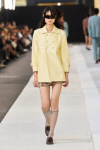 Model walking a runway in a pale yellow double-breasted coat, short metallic-looking skirt, beige knee-high socks, brown loafers, and oversized black visor sunglasses.