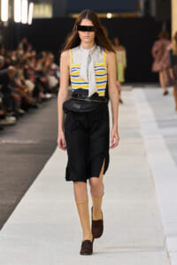 Model walks a runway in a yellow and blue striped bustier top, white scarf, black high-waisted shorts with a waist bag, and tan socks with brown shoes.