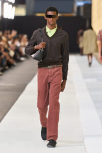 Male model walks a runway in a dark V-neck sweater over a lime-green patterned shirt, red pants, black belt, and a black shoulder bag with futuristic sunglasses.