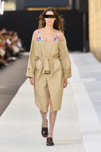 Model walking a fashion show in a tan belted trench with sheer sleeves over a colorful patterned top and a black visor-style sunglasses.
