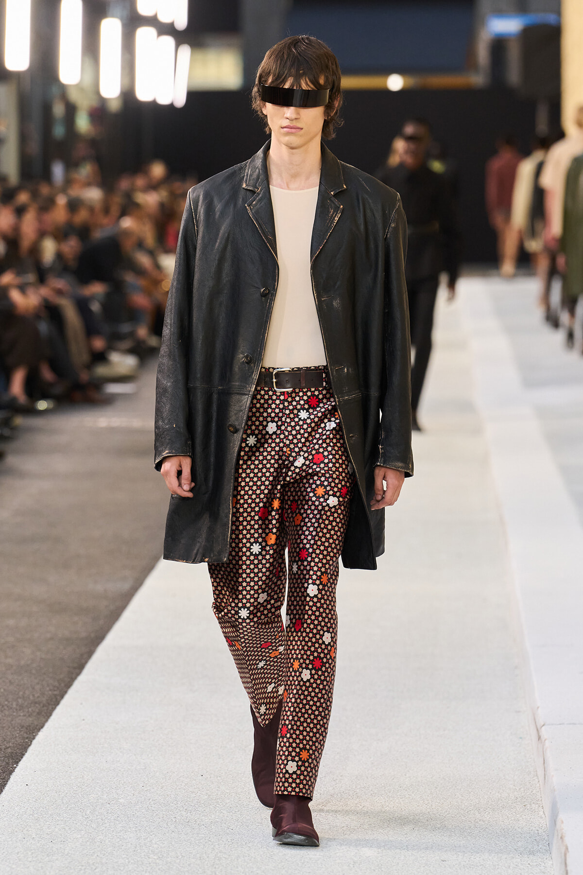 Runway model wearing a long black leather coat over a light shirt, patterned floral pants, burgundy boots, and a black eye-visor.