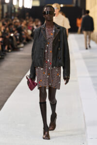 Model on a runway wearing a black leather jacket over a floral printed dress, with a futuristic visor and brown ankle boots, holding a burgundy clutch.