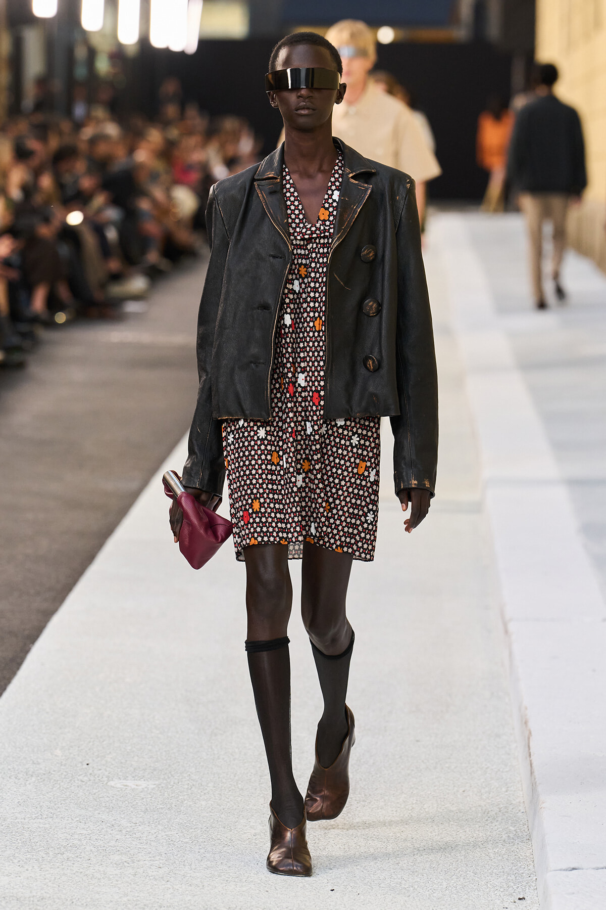 Model on a runway wearing a black leather jacket over a floral printed dress, with a futuristic visor and brown ankle boots, holding a burgundy clutch.