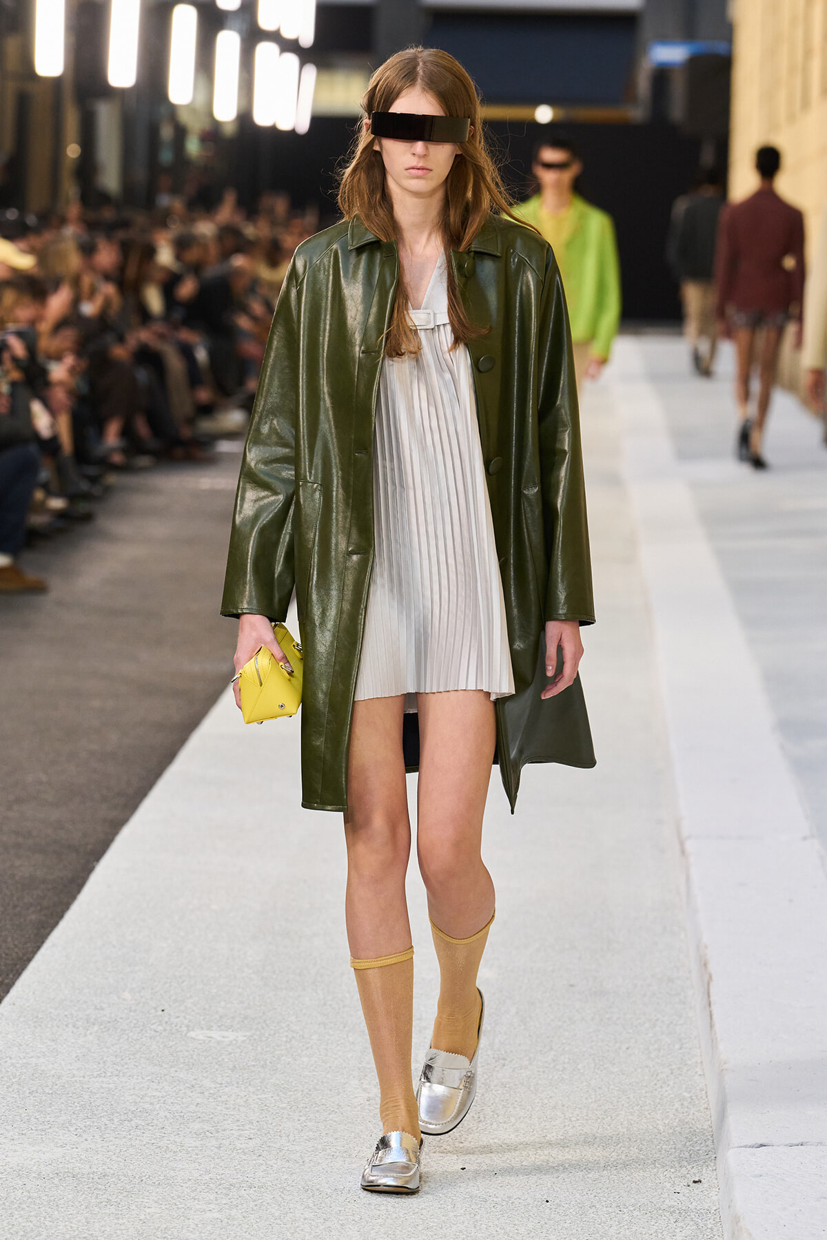 Model walking a runway in a dark green leather coat over a light pleated dress, with a yellow clutch, mustard socks, silver loafers, and a black visor-style sunglasses.