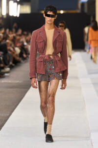 Male model walks the runway in a pink belted jacket, beige top, and floral shorts, with a black blindfold-style eyepiece.