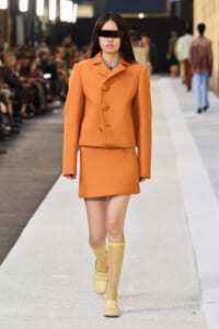 Model walks the runway in an orange tailored coat and matching mini skirt, wearing yellow socks and loafers with a black eyepatch-style accessory over the eyes.