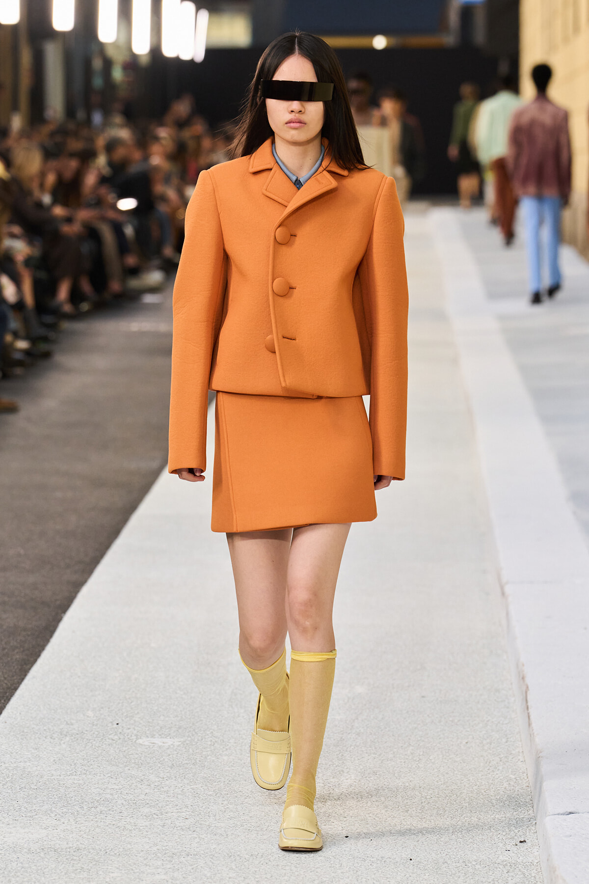 Model walks the runway in an orange tailored coat and matching mini skirt, wearing yellow socks and loafers with a black eyepatch-style accessory over the eyes.