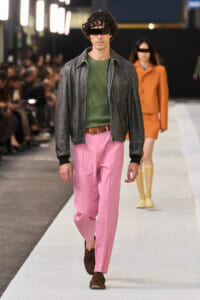 Male model walking runway in green knit sweater, black leather jacket, pink trousers, brown shoes; futuristic black visor across eyes.