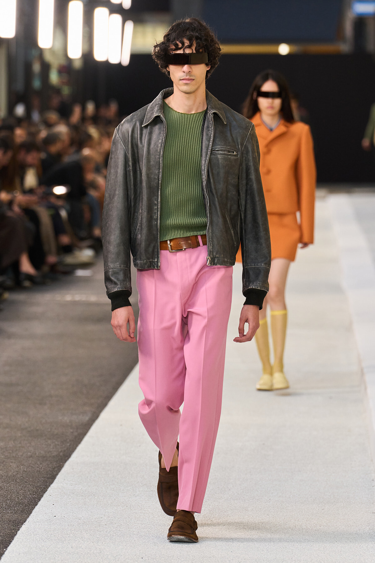 Male model walking runway in green knit sweater, black leather jacket, pink trousers, brown shoes; futuristic black visor across eyes.