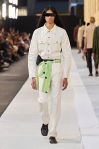 Model walks the runway in an all-white denim-style suit with a bright green floral belt and black rectangular sunglasses, brown loafers.