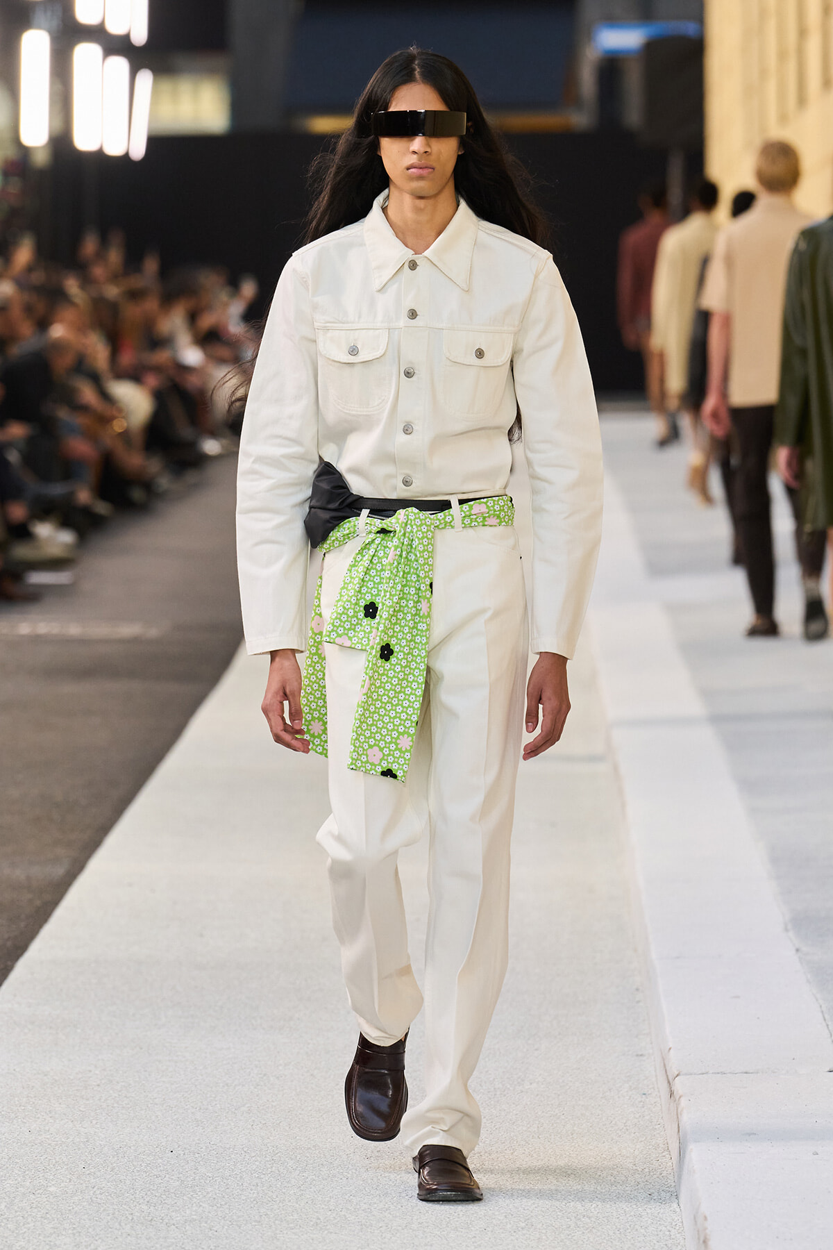 Model walks the runway in an all-white denim-style suit with a bright green floral belt and black rectangular sunglasses, brown loafers.