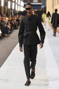 Male model walks a runway in an all-black outfit: structured button-down shirt with shoulder cutouts, slim trousers, belt bag, and futuristic sunglasses.