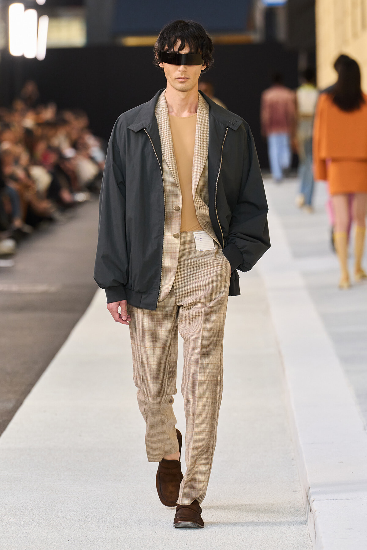 Male model walks a runway in a beige plaid suit with a tan top, layered under a dark windbreaker and black visor sunglasses.
