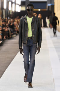 Male model walks the runway in a distressed black leather jacket, lime green patterned shirt, blue jeans, and brown loafers with futuristic sunglasses.