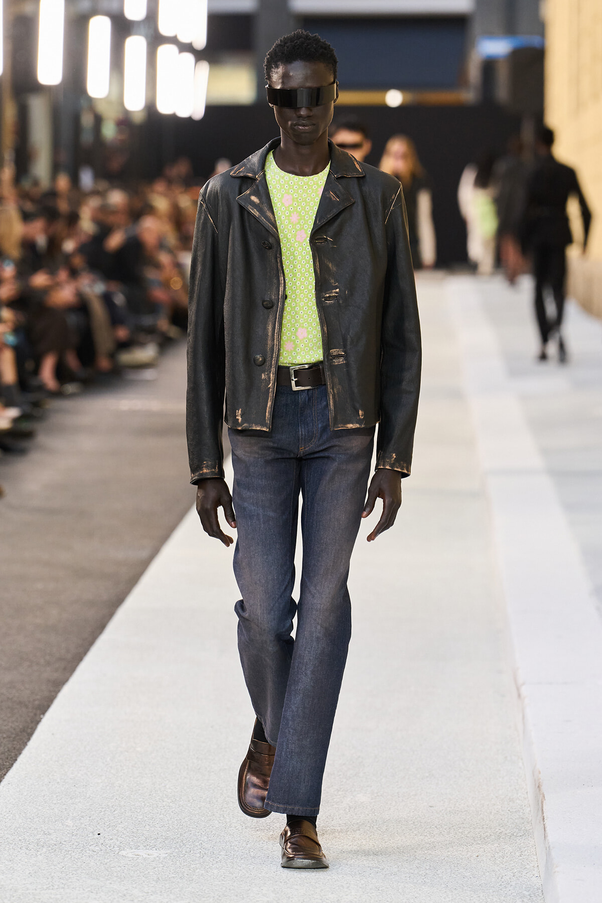 Male model walks the runway in a distressed black leather jacket, lime green patterned shirt, blue jeans, and brown loafers with futuristic sunglasses.