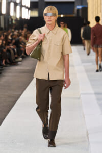 Male model on a runway wearing a beige short-sleeve shirt, brown pants, black shoes, a silver blindfold, and carrying a green bag over the shoulder.