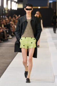 Male model on a runway wearing a black coat over a beige knit top and lime-green floral shorts, with beige socks and black loafers, plus a futuristic visor.
