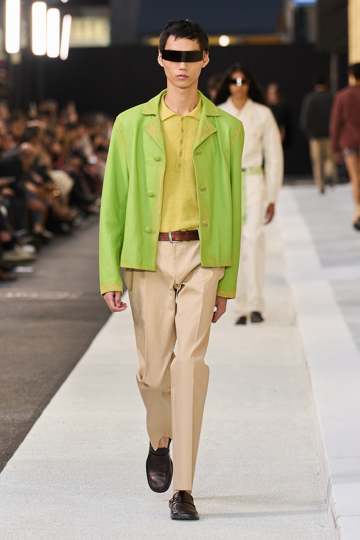 Male model walks a runway wearing a lime-green jacket over a yellow polo and beige trousers with a brown belt and dark visor-style sunglasses.