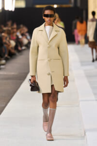 Model walks the runway in a pale beige knee-length coat with oversized lapels and large buttons, wearing futuristic black sunglasses and carrying a small dark clutch.