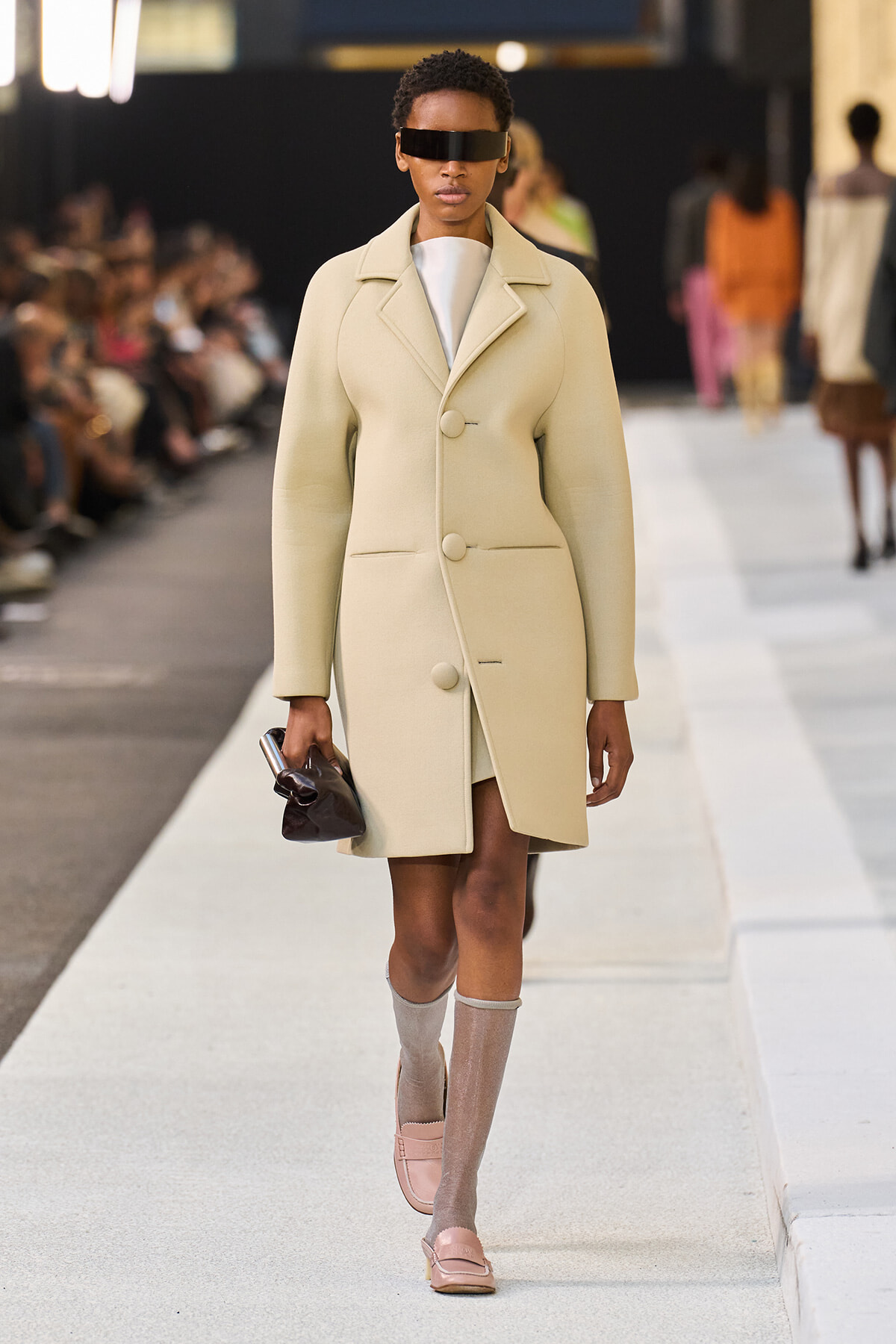 Model walks the runway in a pale beige knee-length coat with oversized lapels and large buttons, wearing futuristic black sunglasses and carrying a small dark clutch.