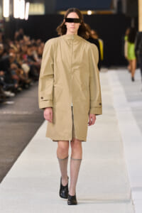 Model walks the runway in a tan oversized zip-front coat with wide sleeves and black rectangular visor sunglasses.