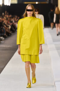 Model on a runway wearing a bright yellow cape-style top with a pleated hem, gold boots, and a black futuristic visor eye shield.