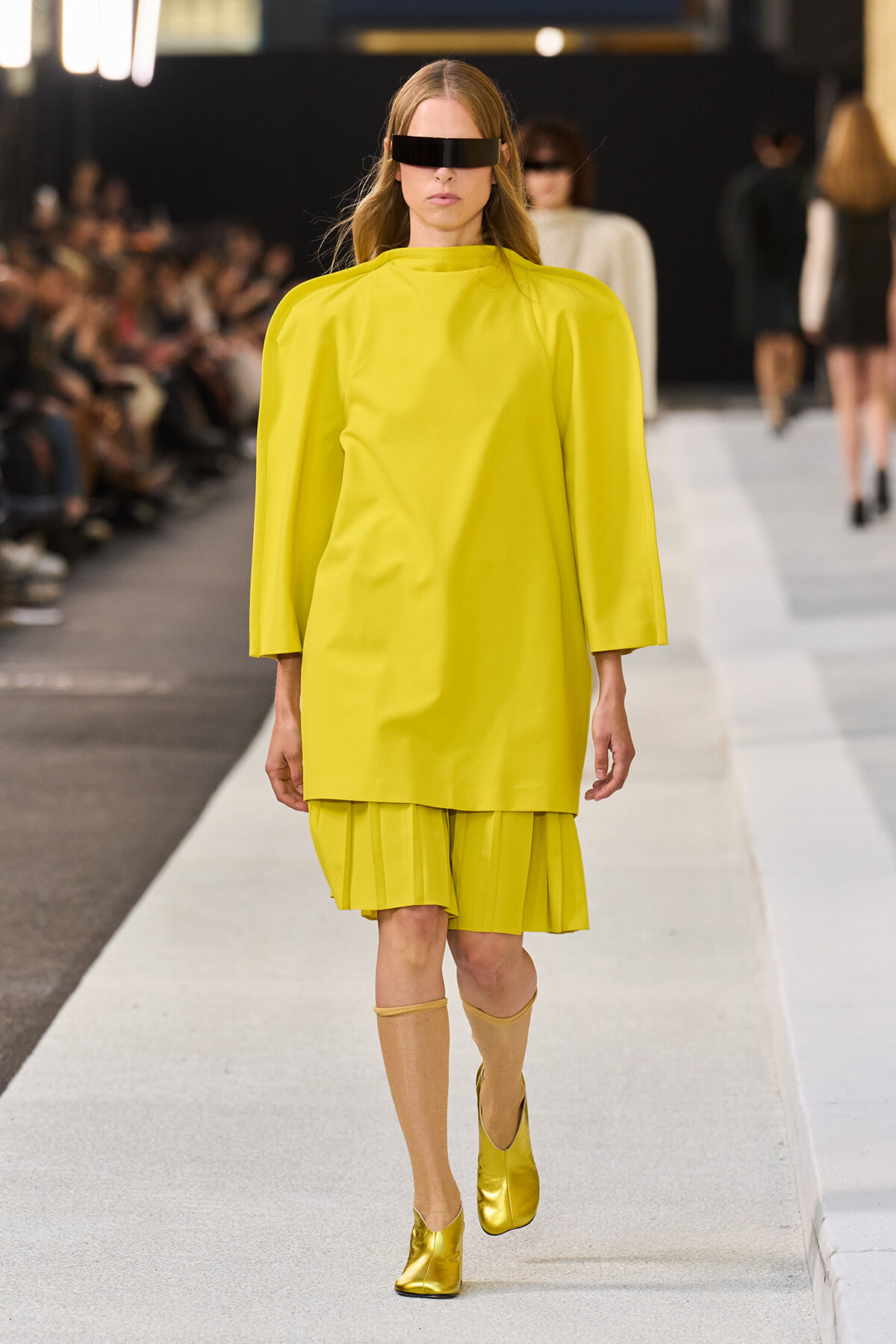 Model on a runway wearing a bright yellow cape-style top with a pleated hem, gold boots, and a black futuristic visor eye shield.