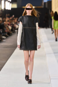 Model walking a fashion runway in a black sleeveless tunic over a beige long-sleeve top, brown shoes, and futuristic visor sunglasses.