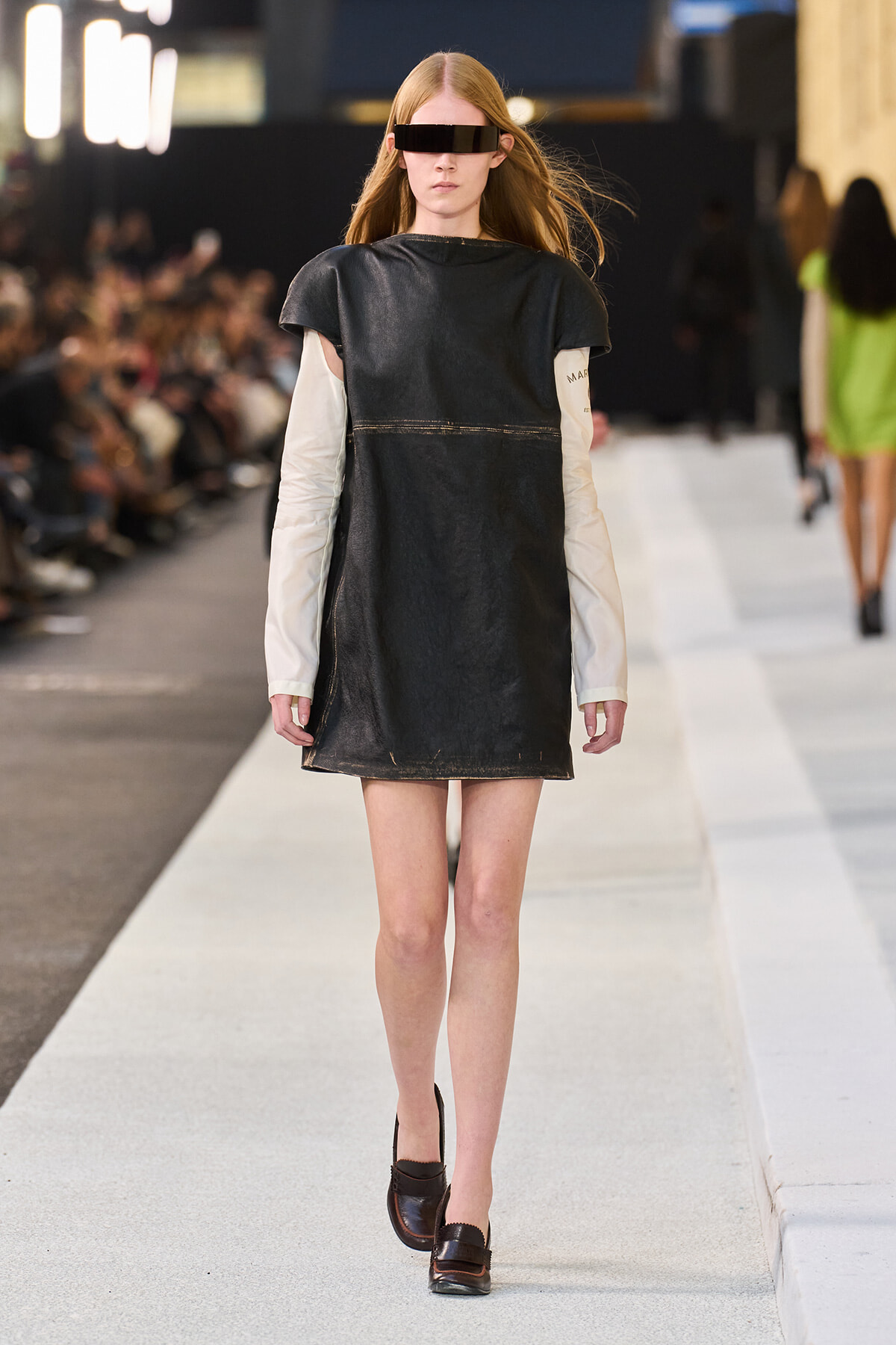 Model walking a fashion runway in a black sleeveless tunic over a beige long-sleeve top, brown shoes, and futuristic visor sunglasses.