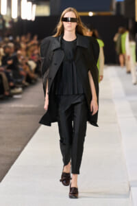 Female model walks a runway in an all-black ensemble with a cape-like coat, pleated top, tailored pants, brown loafers, and futuristic visor sunglasses.