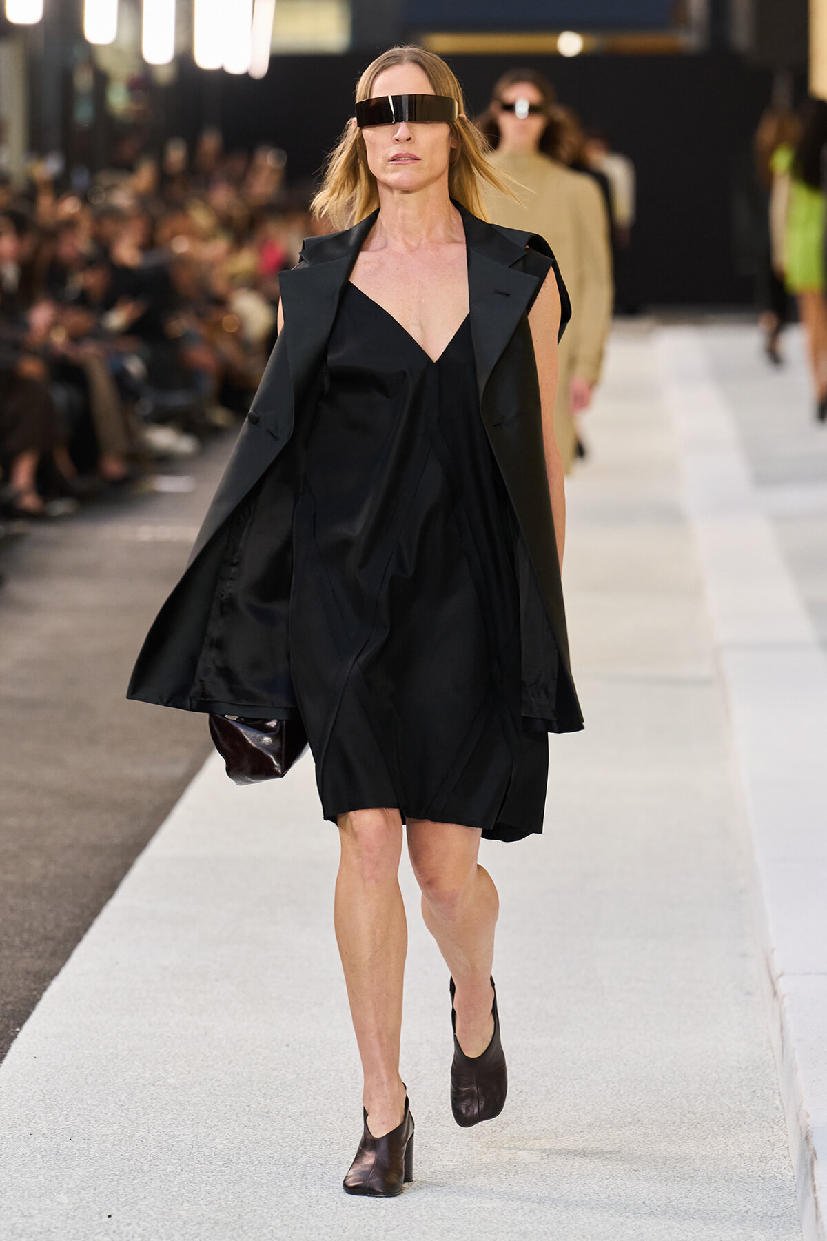 Model walks a runway in a black V-neck dress with a cape-like overcoat and a futuristic wide visor, black ankle boots.