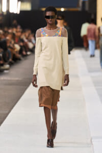 Model walks a fashion runway in a cream off-shoulder top with sheer yoke and colorful patterned straps, paired with brown shorts and dark visor sunglasses.