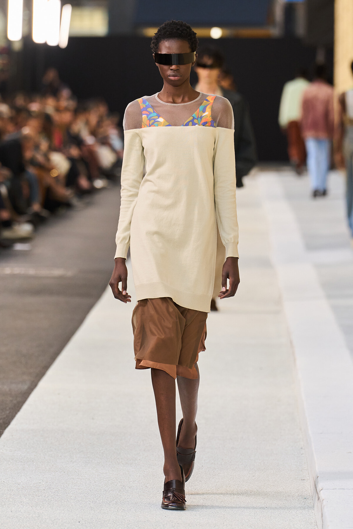 Model walks a fashion runway in a cream off-shoulder top with sheer yoke and colorful patterned straps, paired with brown shorts and dark visor sunglasses.