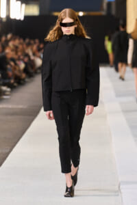 Model on a fashion runway wearing an all-black outfit: zip-up jacket with puffed sleeves and cropped pants, plus a rectangular black visor.