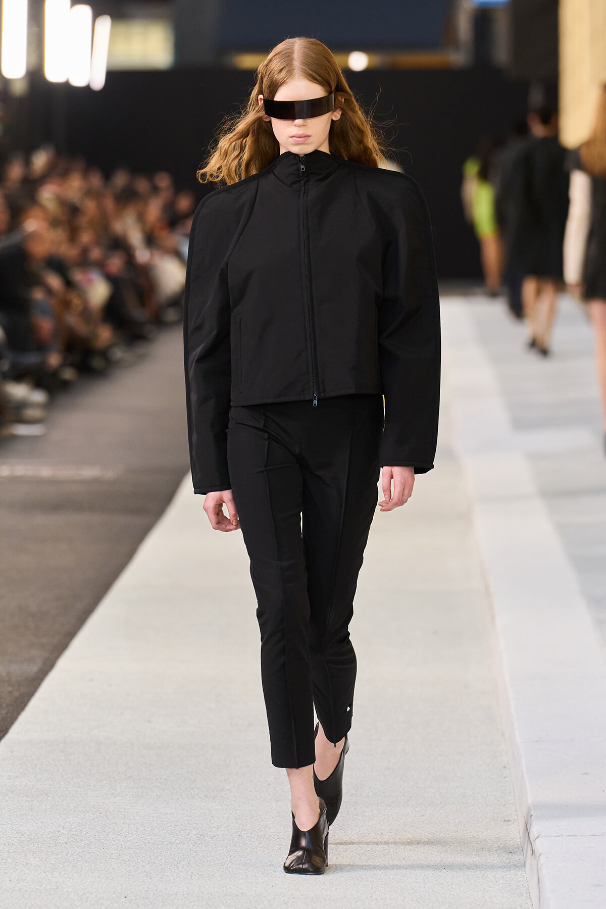 Model on a fashion runway wearing an all-black outfit: zip-up jacket with puffed sleeves and cropped pants, plus a rectangular black visor.